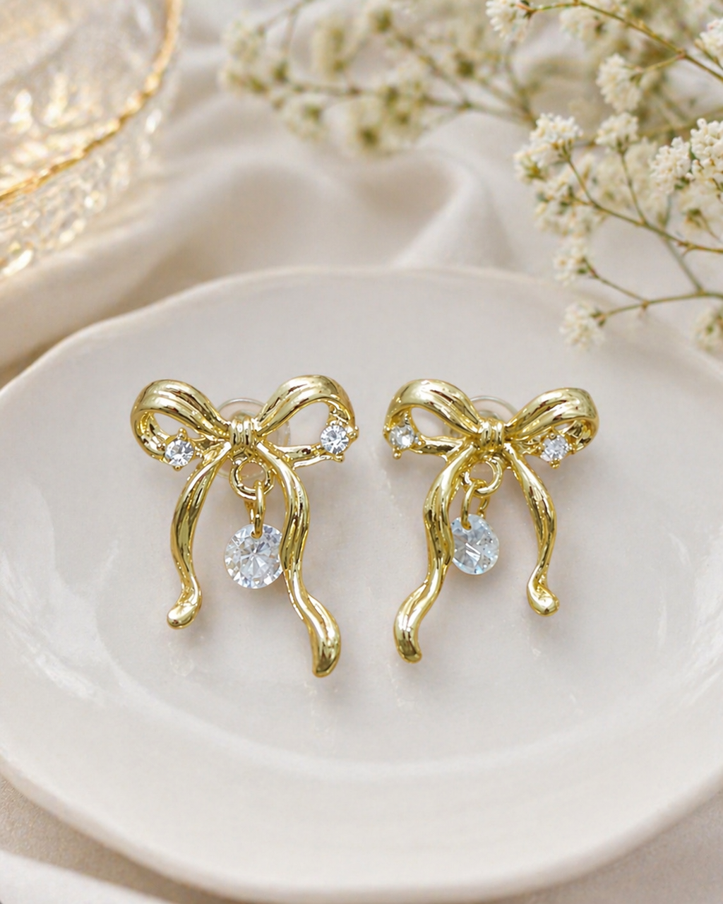 Gold bow-shaped earrings with clear stones on a white dish with dried flowers.