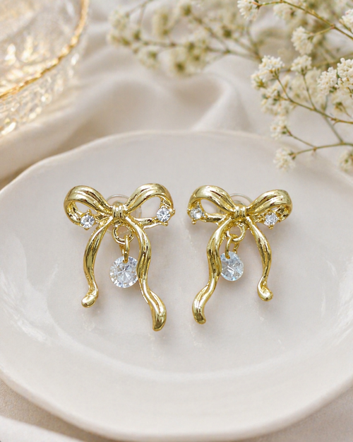 Gold bow-shaped earrings with clear stones on a white dish with dried flowers.